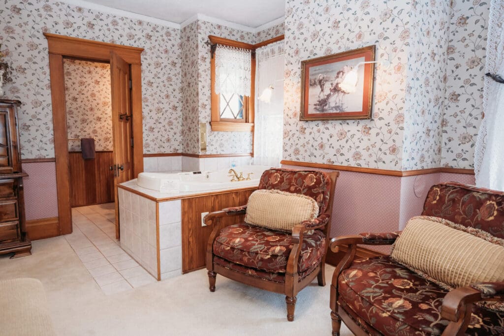 A cozy room in Alexandria with floral wallpaper features two upholstered armchairs, a large bathtub with gold fixtures, a wooden door leading to another area, and lace-curtained windows—perfect for a romantic Valentine's Day retreat. A framed picture hangs above the tub.