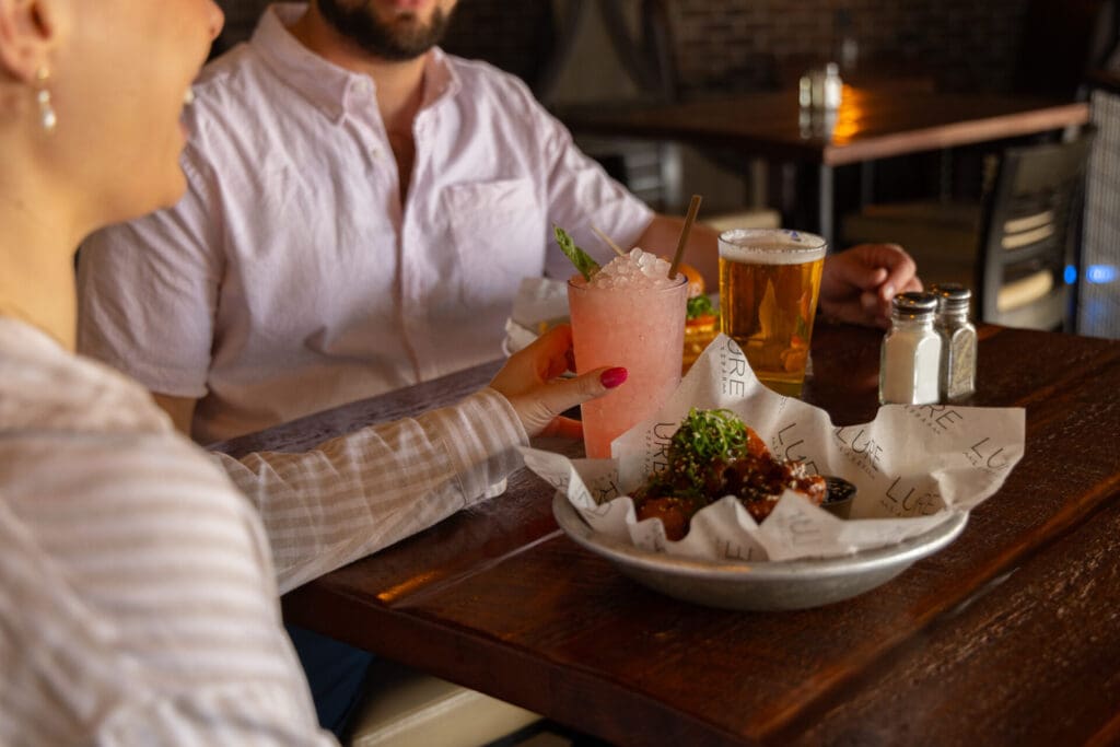 Two people sit at a wooden table in Alexandria with drinks—a pink cocktail and a beer—plus saucy chicken wings garnished with greens. One reaches for their drink, both enjoying a cozy Valentine's Day meal together.
