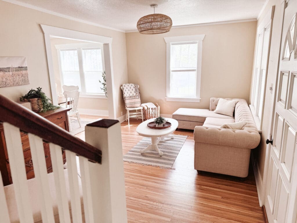 Bright living room with a beige sectional sofa, round white coffee table, wood floors, and neutral decor—perfect for a romantic getaway in Alexandria. Sunlight streams through large windows; a dining area with a white table and chairs is visible behind.