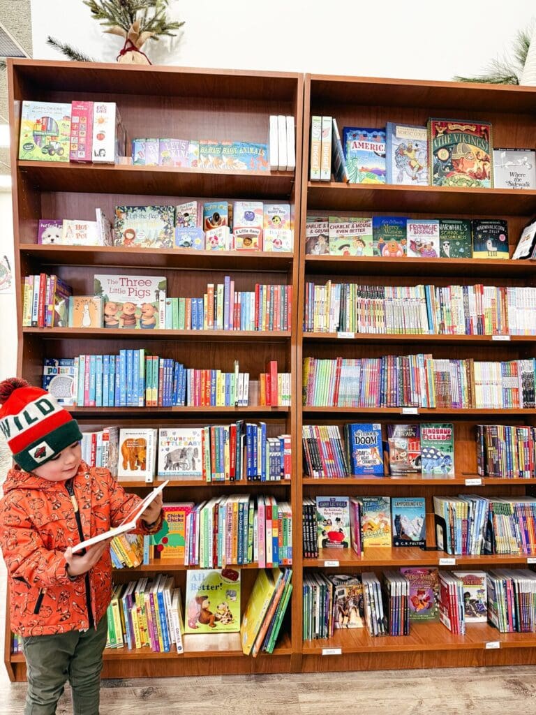 A young child in an orange jacket and winter hat stands in front of tall bookshelves filled with colorful children’s books, enjoying an open book during Saturday Story Hour.