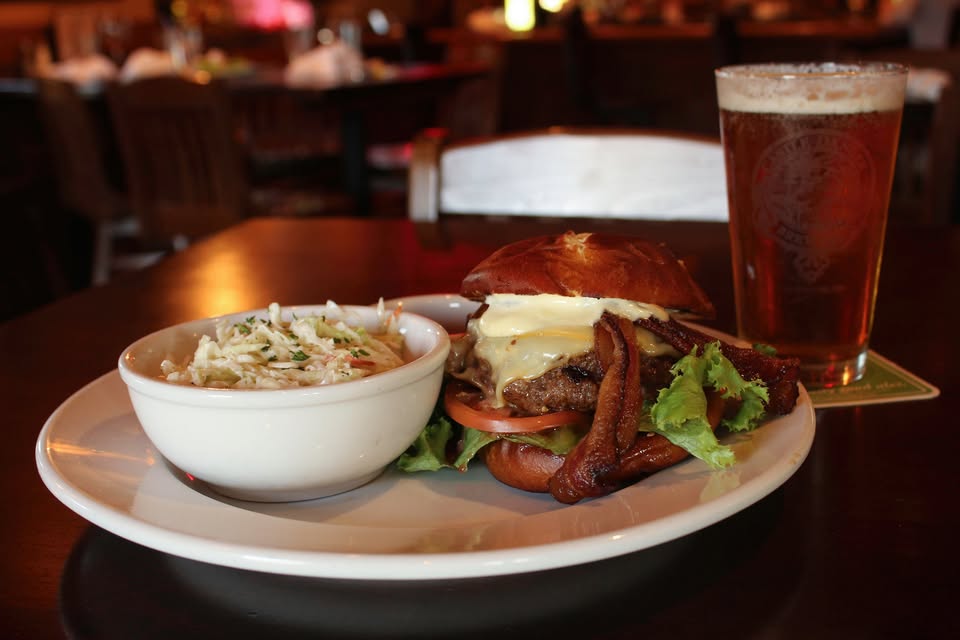 A plate with a cheeseburger topped with bacon, lettuce, and tomato, served with coleslaw. A glass of beer sits beside it on a dark wooden table in an Alexandria restaurant—perfect for a casual Valentine’s Day meal.