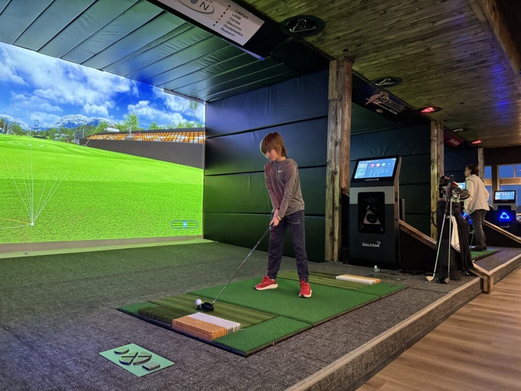 A child stands on an indoor golf simulator mat in Alexandria MN, preparing to swing a club at a digital green course—a perfect Spring Break activity. The area features advanced golf technology, with another person visible in the background.