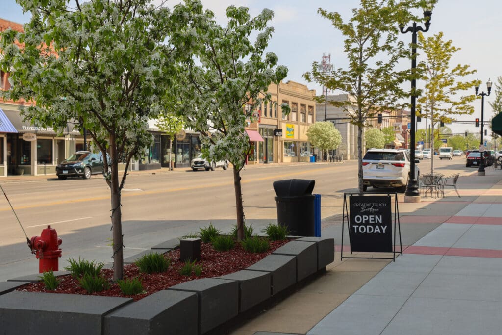 A quiet downtown street in Alexandria MN with small trees, a red fire hydrant, benches, parked cars, and a sidewalk sign that reads "OPEN TODAY" near local shops and buildings—perfect for your Spring Break travel guide on a sunny day.