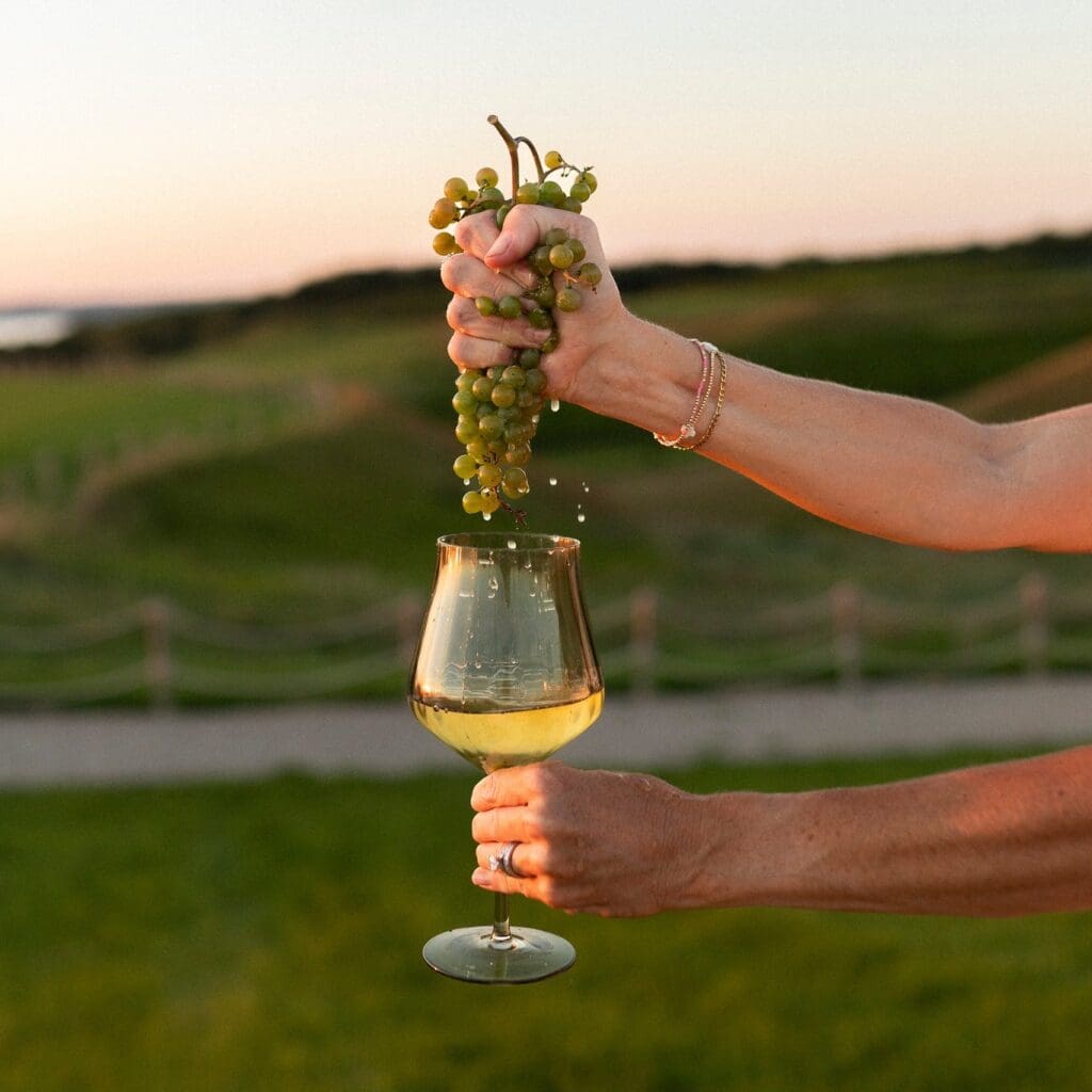 A person squeezes a bunch of green grapes over a wine glass outdoors at Rolling Forks Vineyard, letting the juice drip into the glass with a scenic green landscape in the background at sunset.