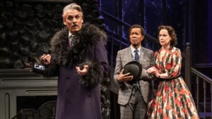 Three actors on stage in a dramatic scene from Agatha Christie's classic mystery play, The Mousetrap: one in a purple fur-collared coat gestures as the others—concerned—stand behind, the man holding a hat and the woman in a floral dress clutching his arm.