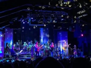 A band performs on a stage lit with blue and purple lights, surrounded by star and moon decorations—an atmosphere that feels drawn from the Collection Unconscious—as musicians play guitars, keyboards, and drums for an attentive audience.