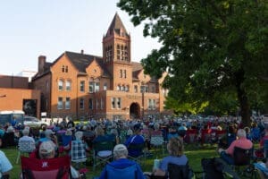 A large crowd sits on lawn chairs facing a historic red-brick building with a clock tower, enjoying the Summer Concert Series performance on the steps, surrounded by trees on a sunny day.