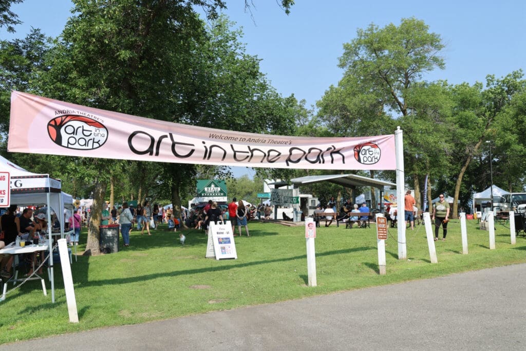A large banner reads “Art in the Park” at the 2026 art festival, where people stroll past tents and vibrant art displays. Green trees and grassy areas fill the background on a sunny day.