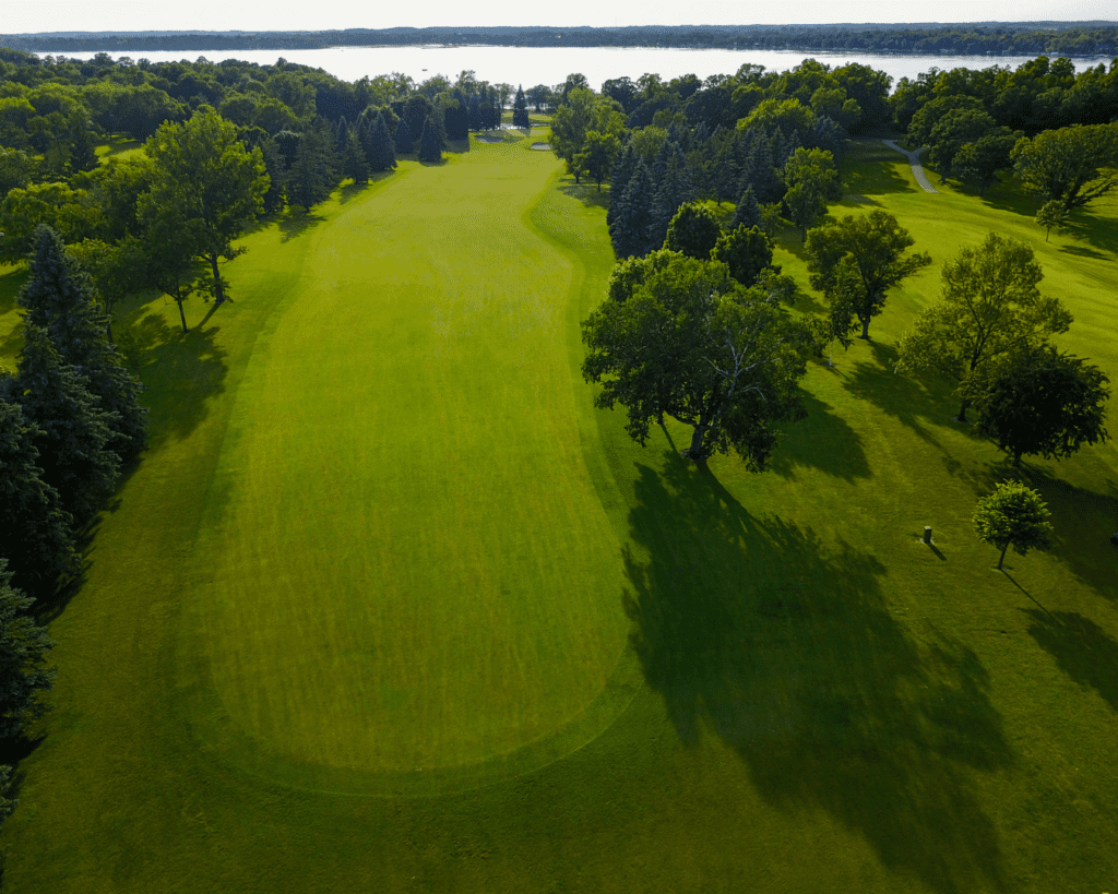 Aerial view of a lush green golf course fairway in Alexandria, surrounded by tall trees, with a river or lake in the background under a clear sky—perfect for Memorial Day Weekend.
