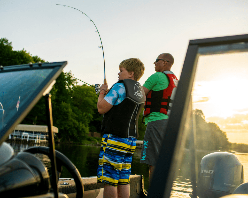 A boy and a man wearing life jackets stand on a boat at sunset during Memorial Day Weekend in Alexandria, with the boy holding a bent fishing rod as if catching a fish. Trees and a dock are visible in the background.