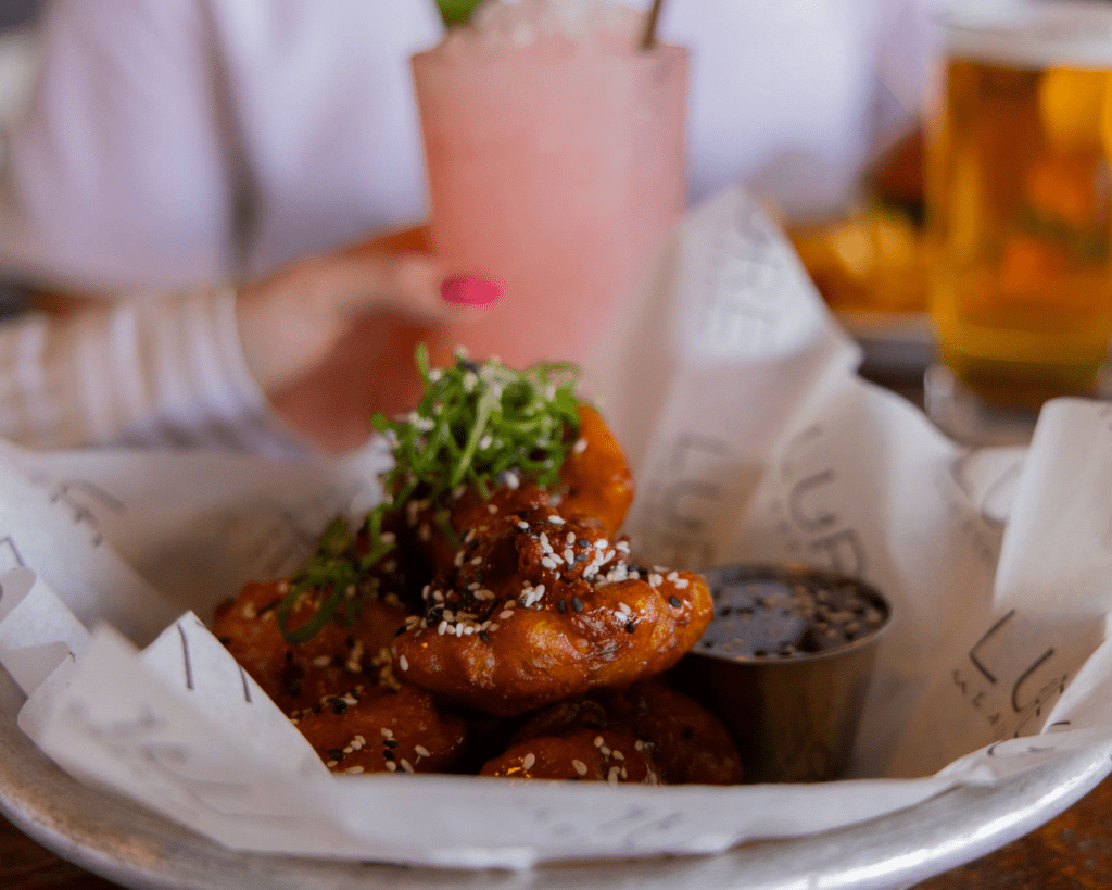 A close-up of saucy chicken wings garnished with sesame seeds and green onions, served in a lined basket with a side of dipping sauce—perfect for sharing during Memorial Day Weekend in Alexandria. In the background, a person holds a pink drink.