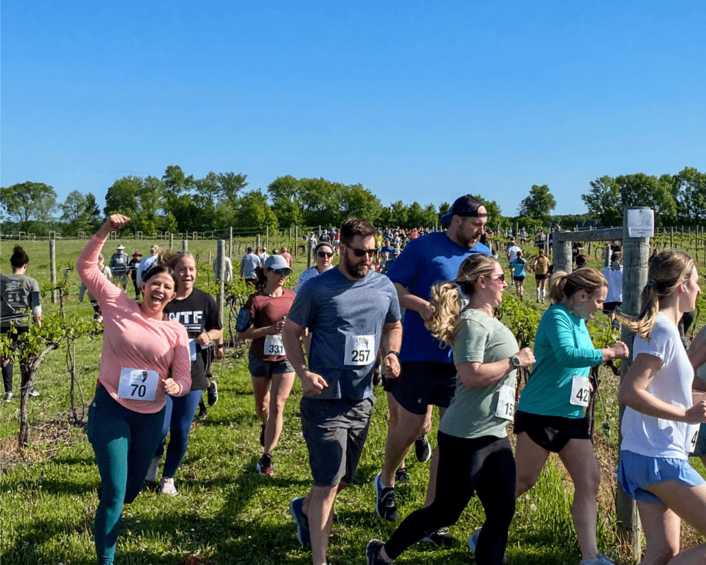 A group of runners participate in an outdoor Memorial Day race on a sunny day in Alexandria, moving along a grassy path surrounded by greenery. One woman in front is smiling and raising her fist in excitement, while others run or walk nearby.