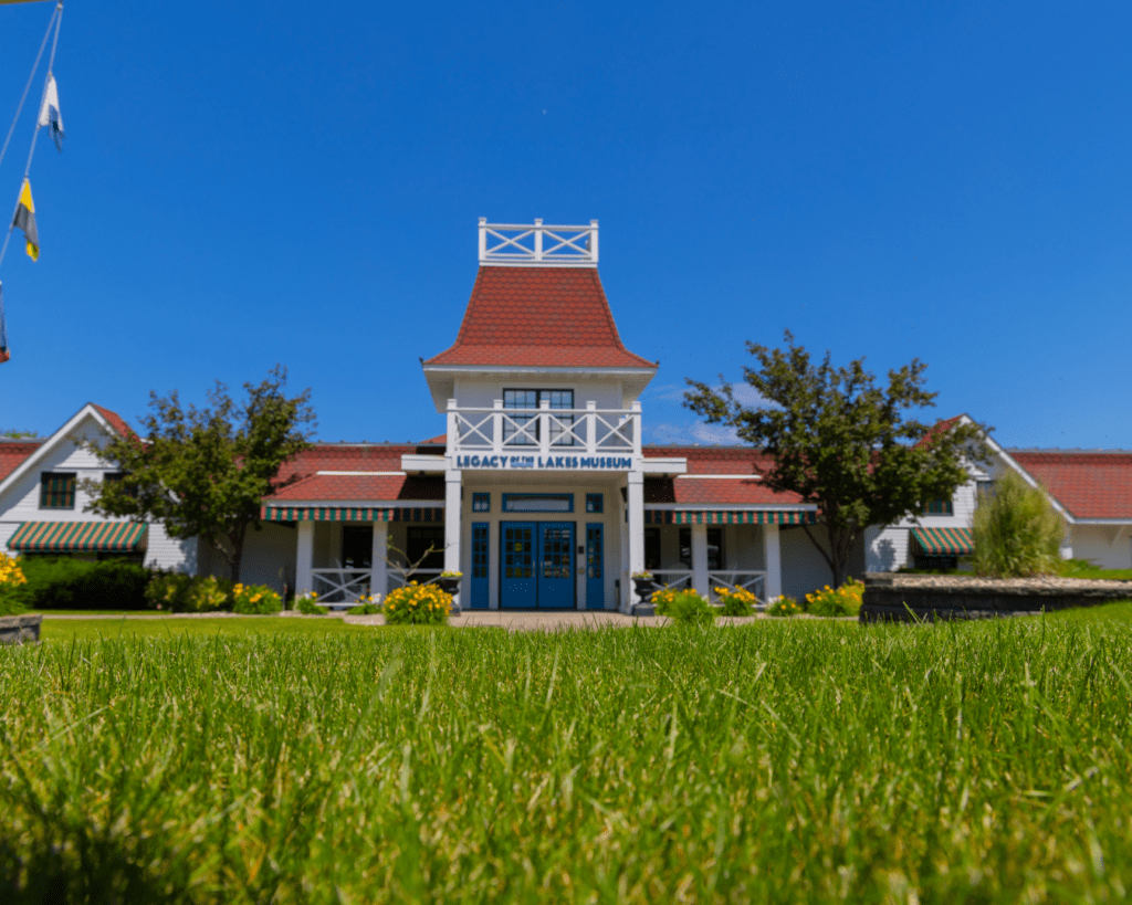 A white building with a red roof and blue doors, labeled "Lakes Museum," sits under a clear blue sky in Alexandria, surrounded by green grass, yellow flowers, and trees—perfect for a Memorial Day Weekend visit.