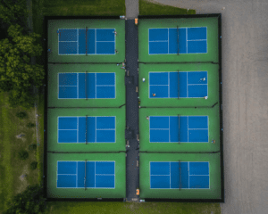 Aerial view of six blue and green tennis courts arranged in two rows of three, with several people playing or standing on the courts. Grass and paths surround the courts on two sides.