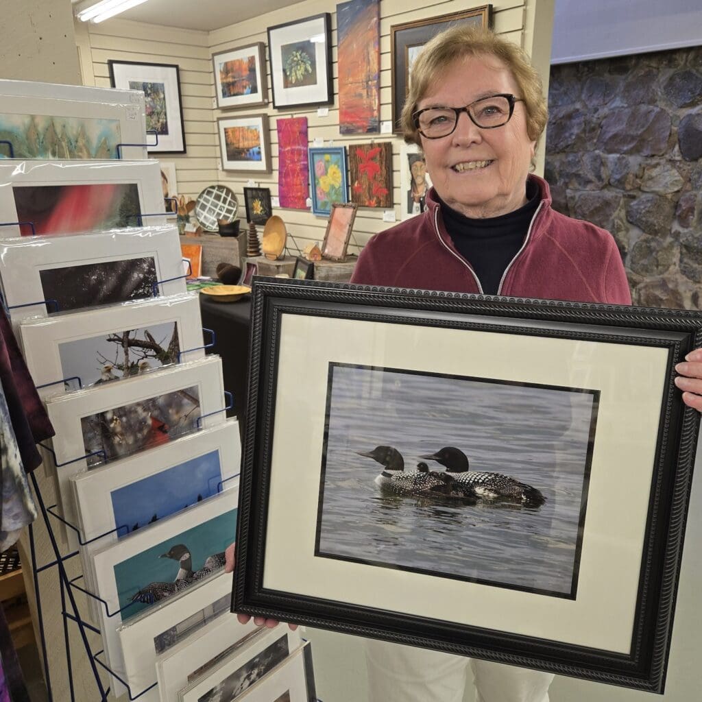 A smiling woman with glasses holds a framed nature photography print of two loons on the water, standing in an art shop surrounded by various framed artworks and photo cards, celebrating Photography Day.