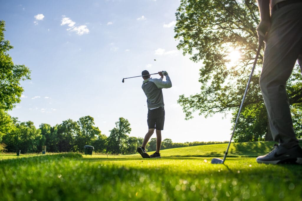A person swings a golf club on a sunny, green Lake Country golf course, while another stands nearby holding a club. Trees and blue sky with scattered clouds are visible in the background—perfect for your Summer Bucket List.