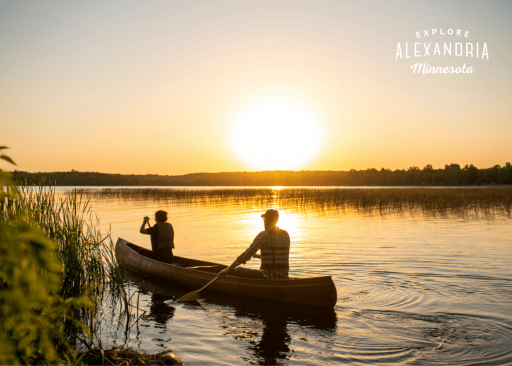 Two people paddle a canoe on a calm lake at sunset near Alexandria, surrounded by reeds and trees, with text in the sky that reads, "Explore Alexandria Minnesota—perfect for nature lovers and trap shooters alike.