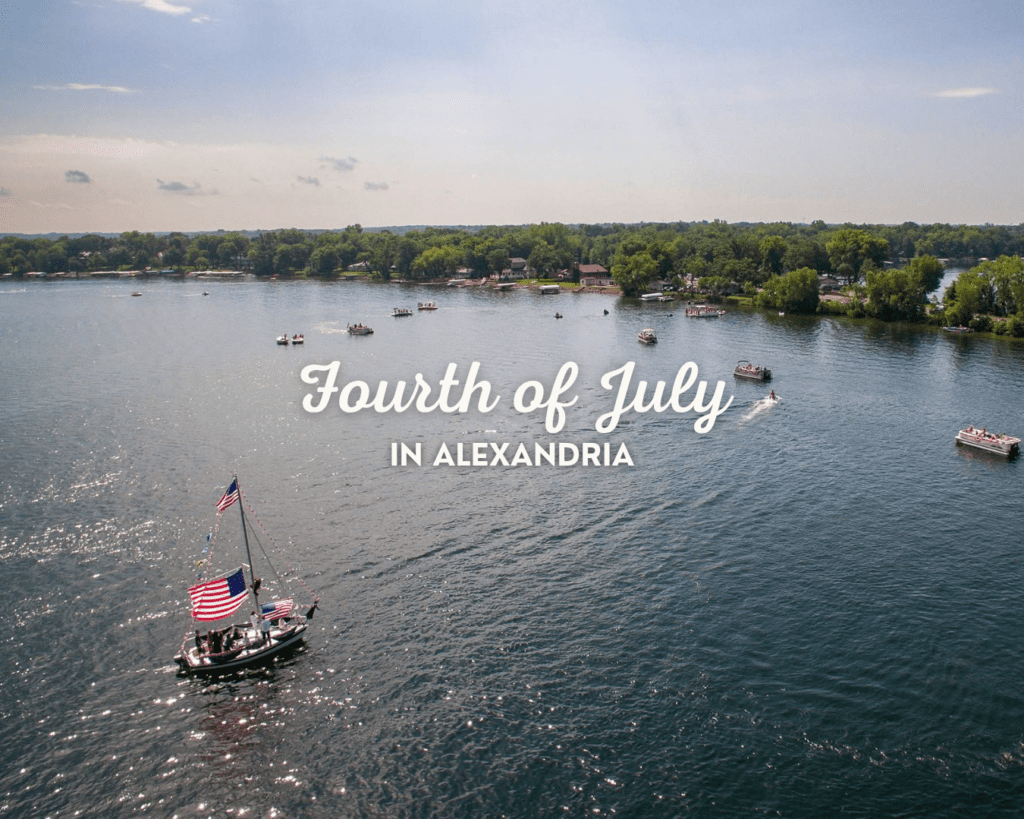 A small boat decorated with American flags floats on a lake in Alexandria, surrounded by other boats and a tree-lined shore. Text reads, "Fourth of July in Alexandria.
