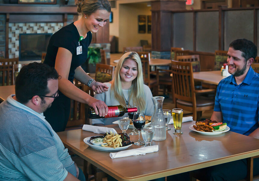 A waitress at Pioneer Grill pours red wine for a smiling man at a restaurant table with two other happy diners, one with long blonde hair and one with a beard. Plates of food and drinks fill the warmly lit setting.