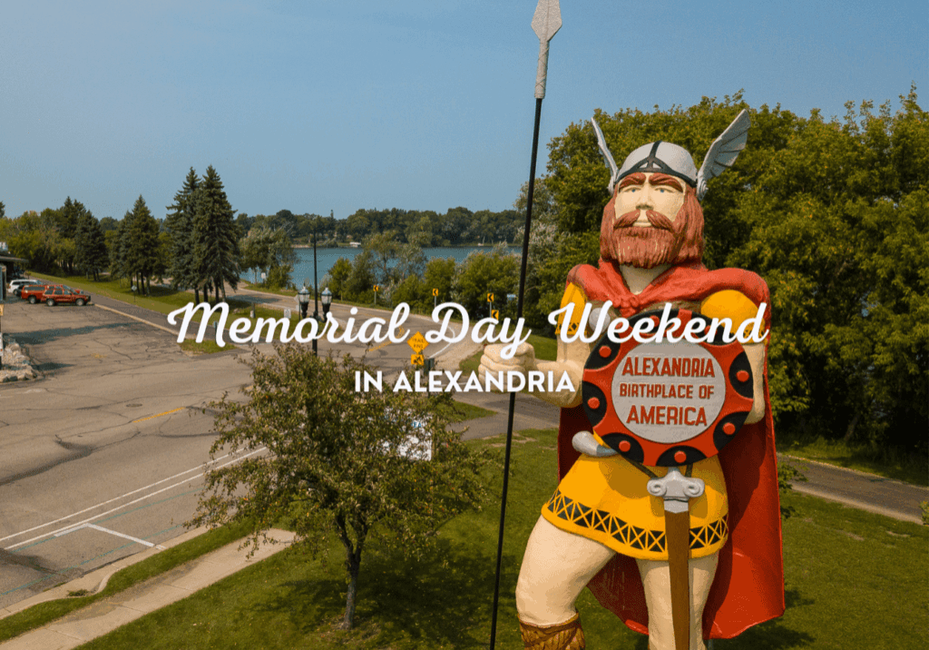 A large Viking statue holds a shield reading "Alexandria Birthplace of America" by the road and green trees, with "Memorial Day Weekend in Alexandria" text across the image. A lake and park are visible in the background, highlighting Memorial Day Weekend.