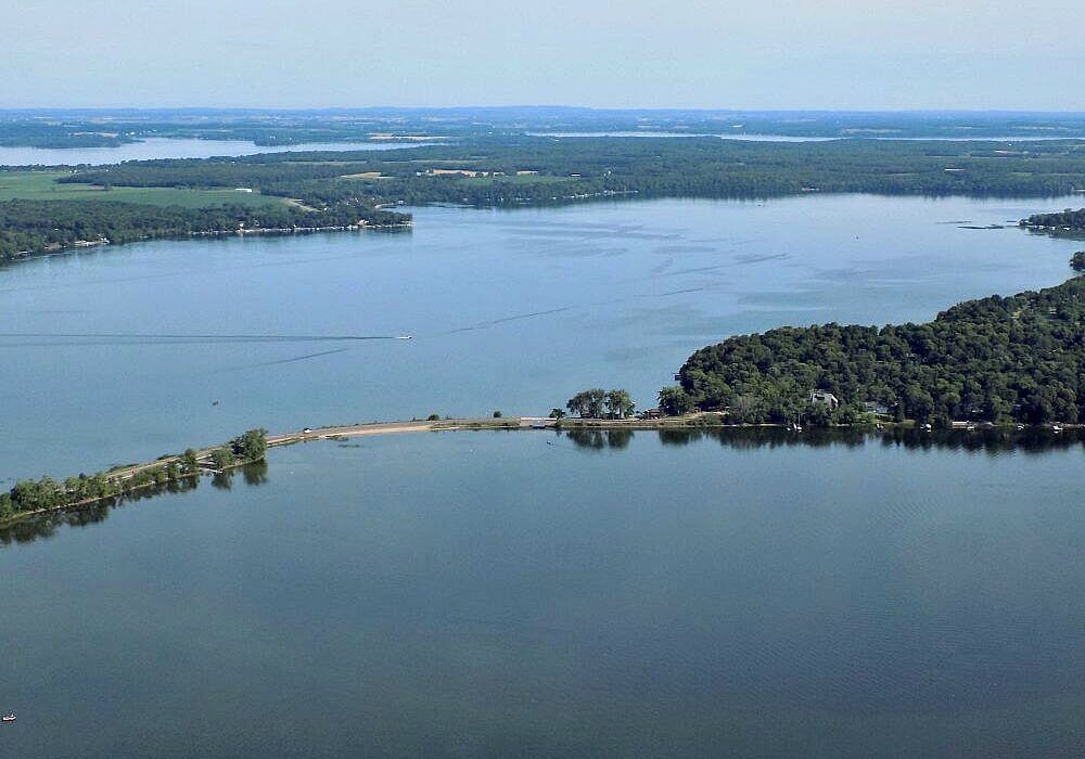 Aerial tour of Alexandria, Minnesota reveals a calm, expansive lake bordered by dense green forest and a narrow strip of land extending into the water under a clear sky.