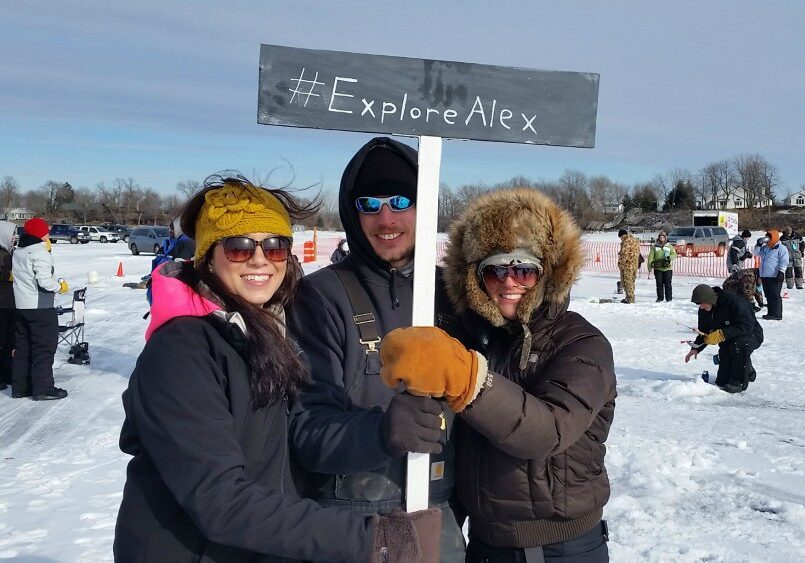 Three people dressed in winter clothes stand on snow holding a sign that reads "#ExploreAlex" near an ice fishing event in Alexandria, MN. Other people are visible in the snowy background, along with orange cones and a fence.
