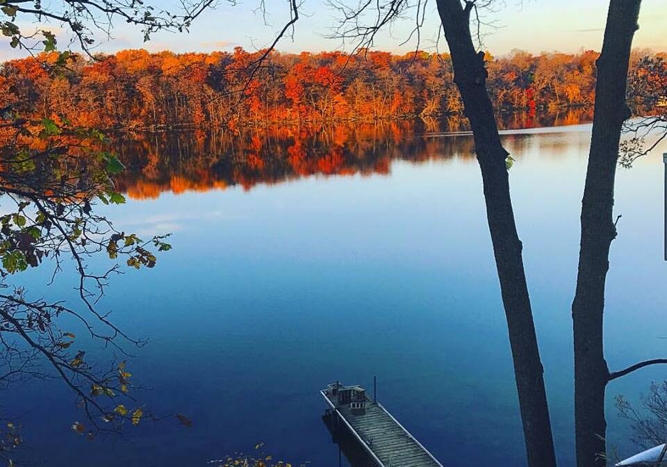 A wooden dock extends into a calm lake, reflecting autumn trees in shades of orange and yellow—a scene perfect for a collection of your fall color photo submissions. Bare branches frame the view under a clear blue sky.