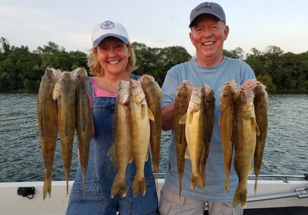 Two smiling people on a boat hold up several large fish each, showcasing their success with late June walleye and sunfish tactics, with a river and green trees in the background under a partly cloudy sky.