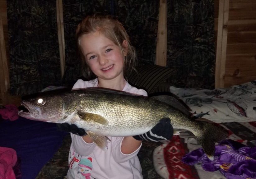 A young girl smiles as she holds a large fish with both hands, wearing gloves—perhaps after an exciting ice fishing adventure—indoors on a bed with patterned blankets.