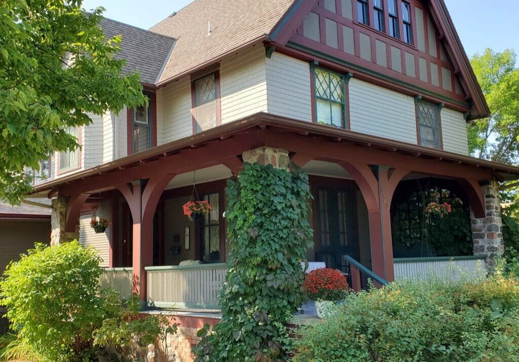 A two-story house with a sloped roof, Tudor-style detailing, and maroon trim, Cedar Rose Inn is a charming bed and breakfast Alexandria gem, featuring a vine-covered porch surrounded by lush greenery and hanging baskets of red flowers.