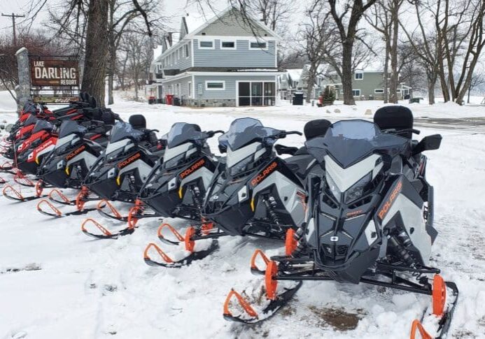 A row of snowmobiles with orange skis, available for rentals at Lake Darling Resort, is parked on snow beside a sign that says "Lake Darling Resort." Snow-covered trees and houses are visible in the background.