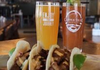 Three tacos filled with meat and garnishes are served on a tray with lime wedges at 205 Grill. Behind them, two drinks in branded glasses sit on a wooden table, creating a cozy food downtown Alexandria Minnesota restaurant scene.