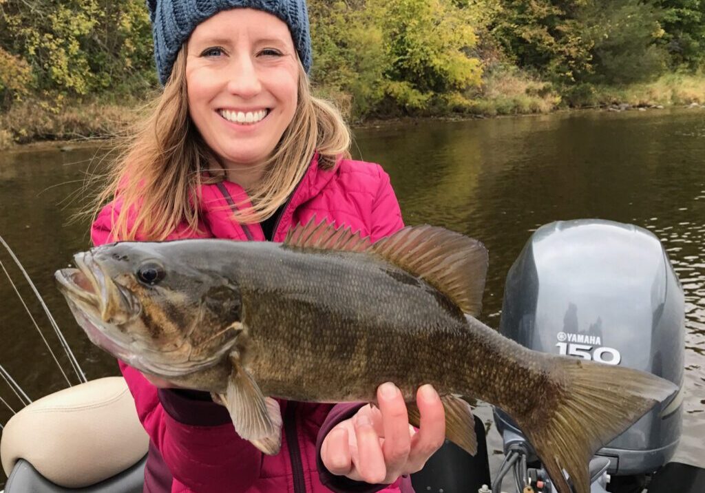 A smiling woman in a red jacket and blue knit hat proudly shows off her fishing catch on a boat, with a river and autumn foliage in the background.