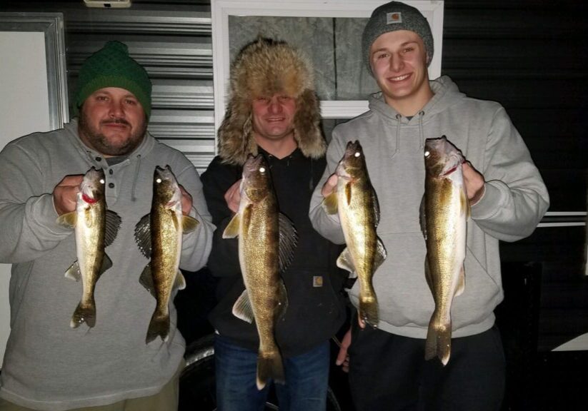 Three men dressed in winter clothing stand indoors, each proudly holding freshly caught fish from their fishing trip, smiling at the camera. The background includes a window and dark paneling.