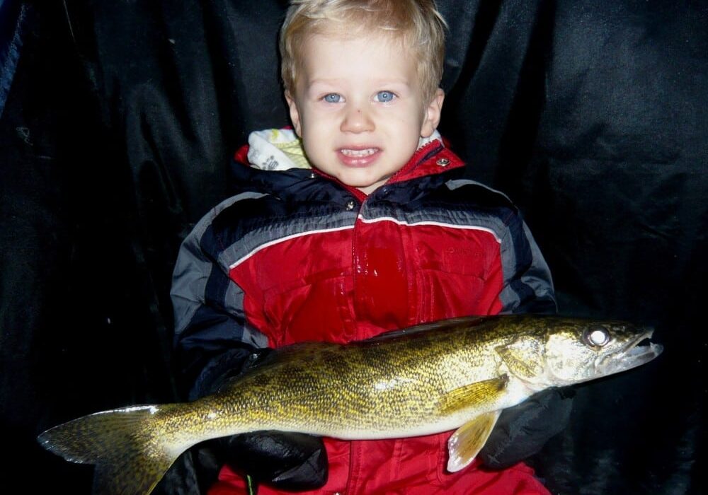 A young child in a red snowsuit smiles while holding a large fish, possibly a walleye, against a dark background—perfect for the new year's ice update and fishing report from Explore Alex.