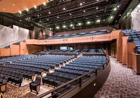 Spacious, modern auditorium at Alexandria Community Education Performing Arts with tiered seating, blue cushioned chairs, and contemporary lighting. Multiple seating levels and patterned carpet complement the large stage area at the front.