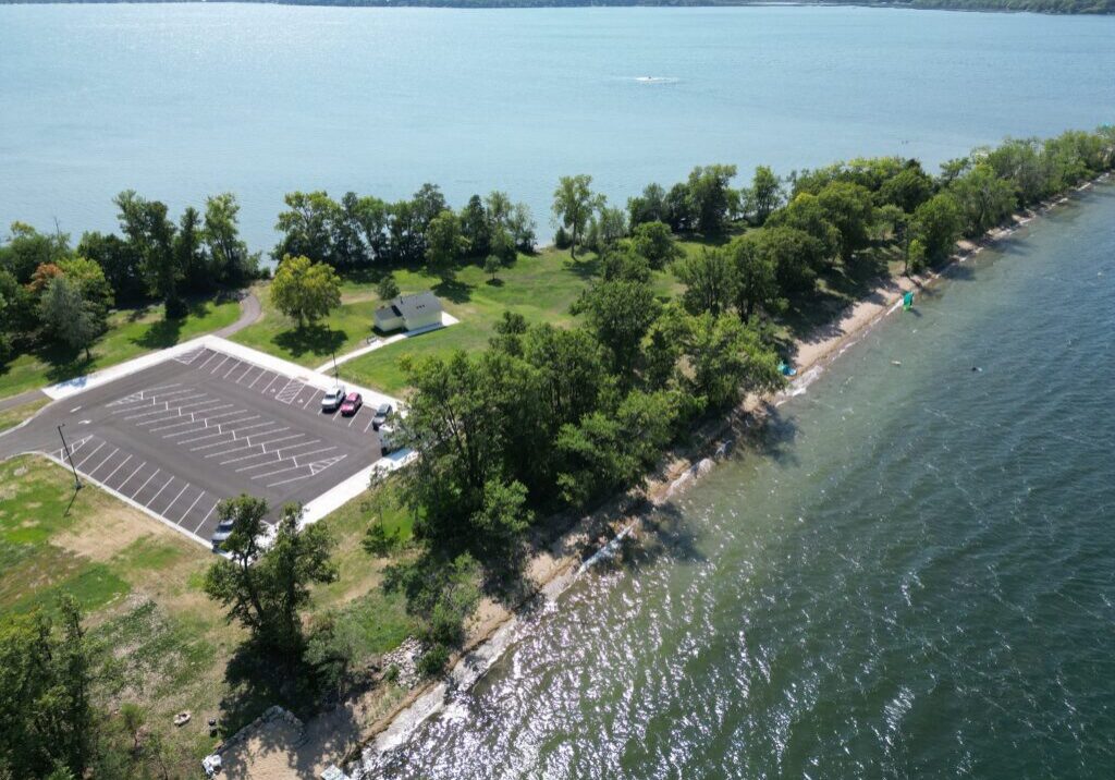 Aerial view of Pilgrim Point County Park with a parking lot, a few cars, lush green trees, and a shoreline where clear blue water stretches into the distance.