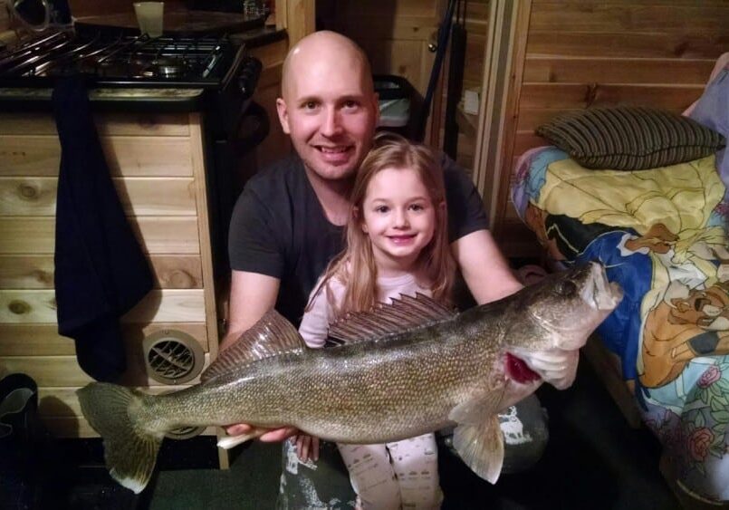 A smiling man and young girl pose indoors, proudly holding a large fish they caught while ice fishing. They are in a cozy wooden room with a bed featuring cartoon character sheets in the background.