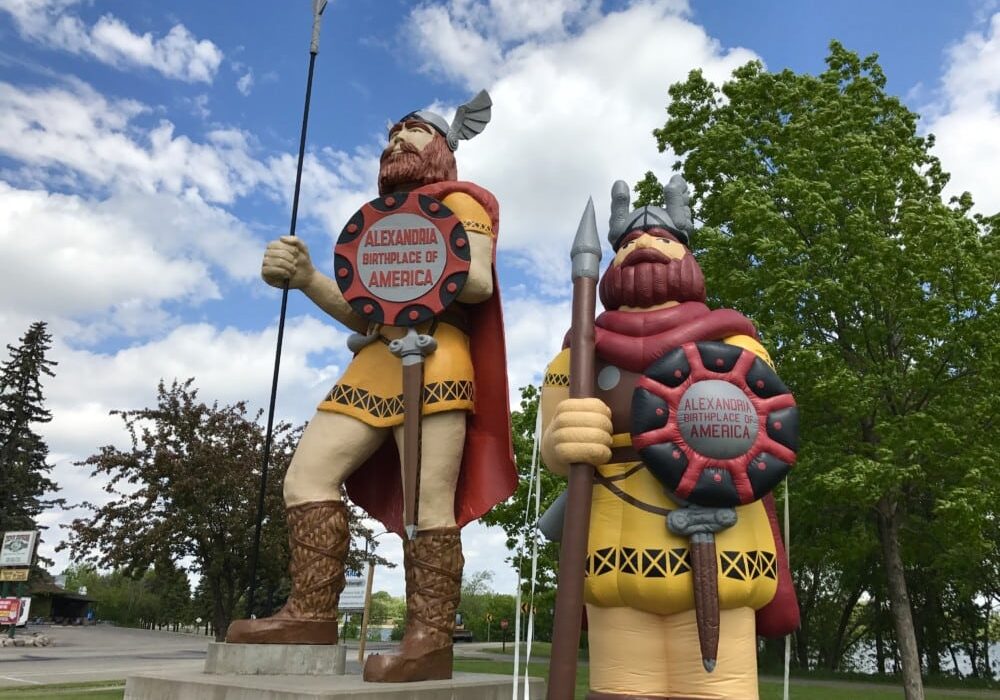 Two large Viking statues with helmets, spears, and shields stand on a concrete platform near green trees. Their shields read "Alexandria, Birthplace of America." Find Little Ole, share your pics and win Alexandria MN tourism prizes!.
