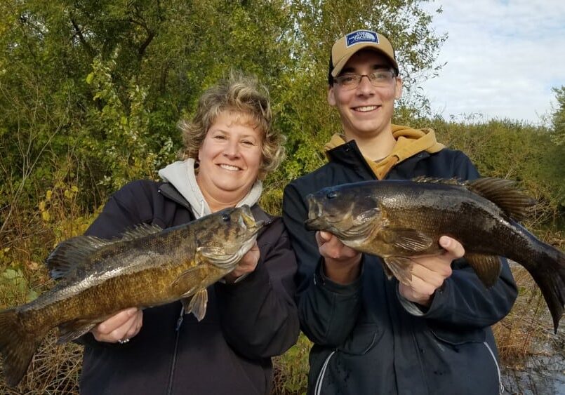 Two people smiling and holding large fish in their hands, standing outdoors by a river or lake with green trees and bushes in the background—a great fishing update at the end of October.
