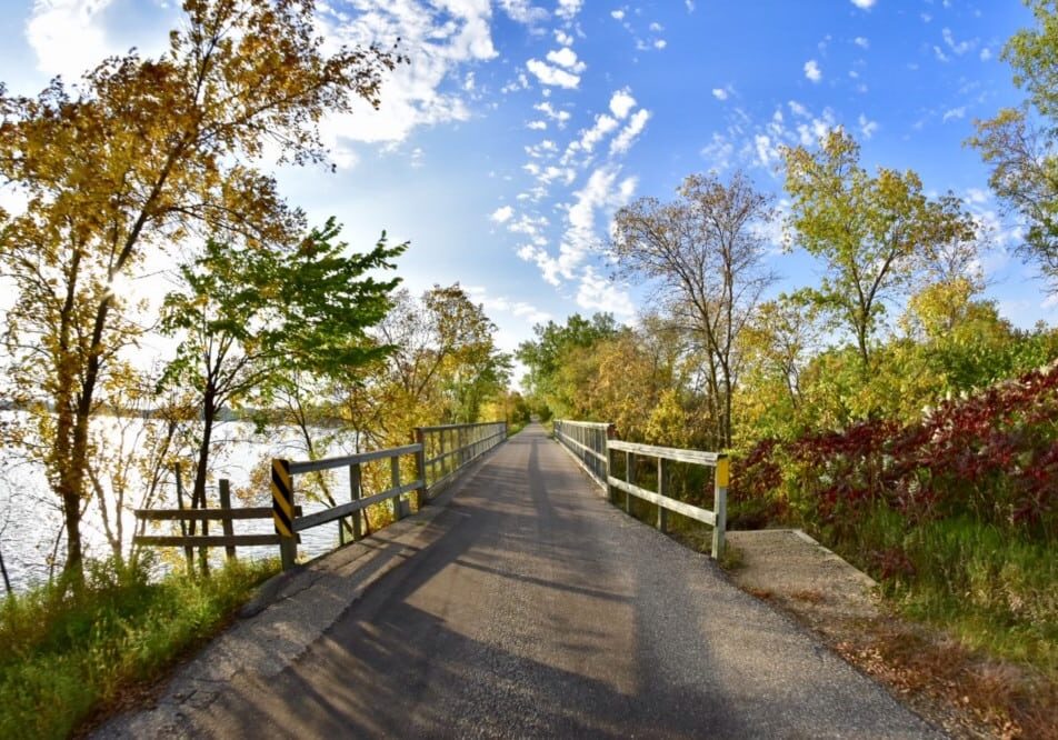 A paved trail with wooden railings winds between trees, their fall colors progressing beautifully in Alexandria Lakes Area, beside a calm body of water under a partly cloudy blue sky.