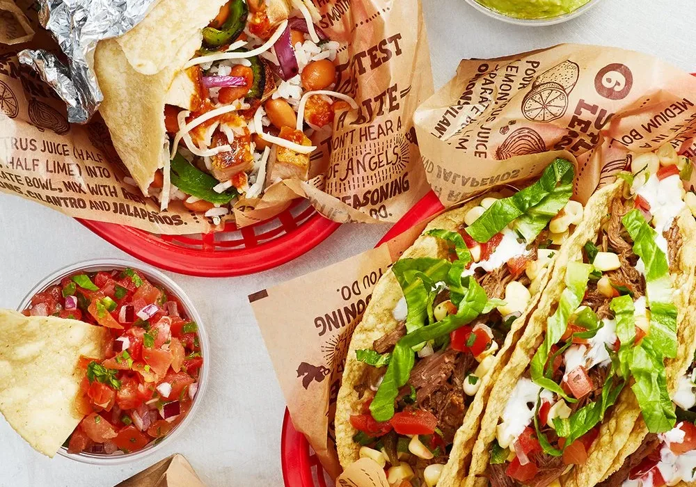 A tabletop at El Loro with a foil-wrapped burrito, two tacos topped with lettuce, tomato, and sour cream, a bowl of guacamole, crunchy tortilla chips, and fresh salsa served on printed paper food trays.