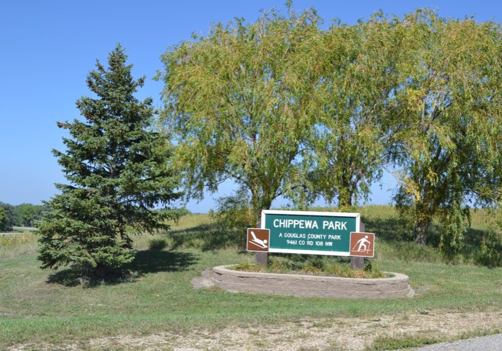 A green sign for Chippewa Park in Minnesota stands on a grassy area beside a pine and a large leafy tree under a clear blue sky. The sign includes icons for boating, swimming, and camping.