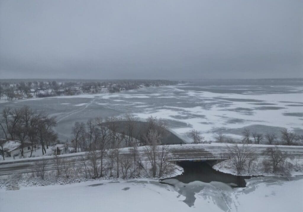 A wintry landscape featuring a partially frozen lake with shimmering ice, snow-covered ground, bare trees, and a small bridge over a creek leading into the lake beneath a gray, cloudy sky.