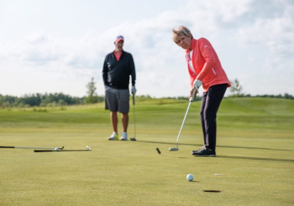 A woman in a coral jacket putts a golf ball toward the hole on a green, aiming to lower your handicap, while a man in a cap and shorts watches her in the background on a sunny day.