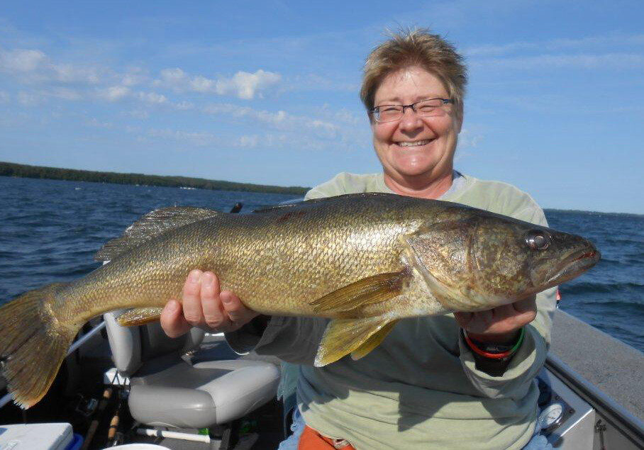 A smiling person wearing glasses holds a large fish on a boat during the 2012 fishing opener, with a lake and blue sky in the background.
