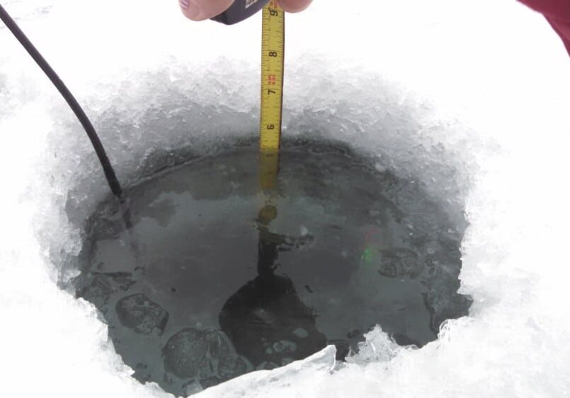 A hand holds a measuring tape in a hole drilled through thick ice on a frozen surface, checking the depth of the water—a typical scene for an Ice Update on current lake conditions in Alexandria Area Lakes.