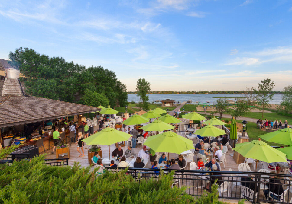 People dine outdoors at the Deck Bar & Grill under bright green umbrellas on a patio overlooking a lake, with trees and a sandy beach in the background on a sunny day.