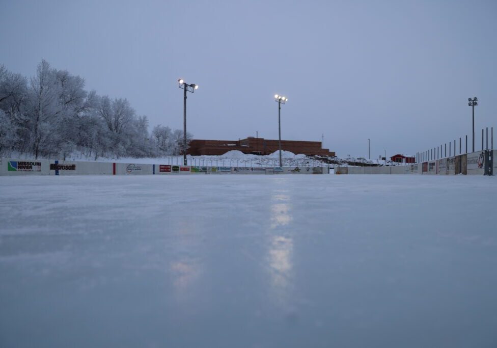 An outdoor ice skating rink at dusk, with snowy trees and Discovery Middle School’s low building in the background. Several tall lights illuminate the rink, reflecting on the smooth ice surface.