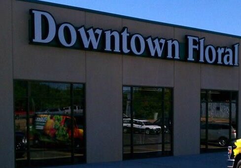 A tan building with large windows displays a bold "Elden's Downtown Floral" sign in white letters. Reflections and vehicles can be seen in the windows and parking lot.